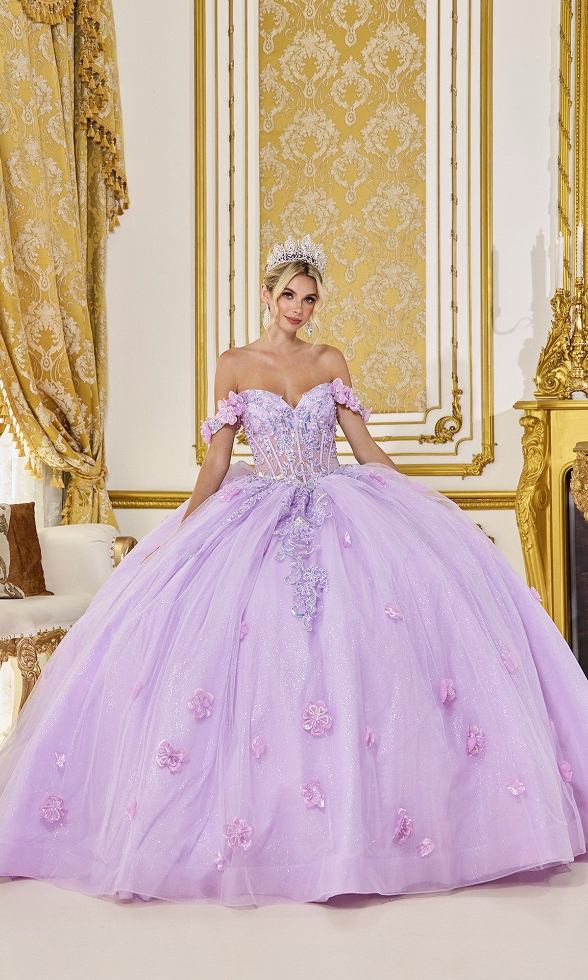A woman in the Quinceanera Dress 5098 by Dancing Queen, a lilac gown with a sheer corset bodice and 3D flower embellishments, stands in an ornate gold-accented room, her look finished with a sparkling tiara.